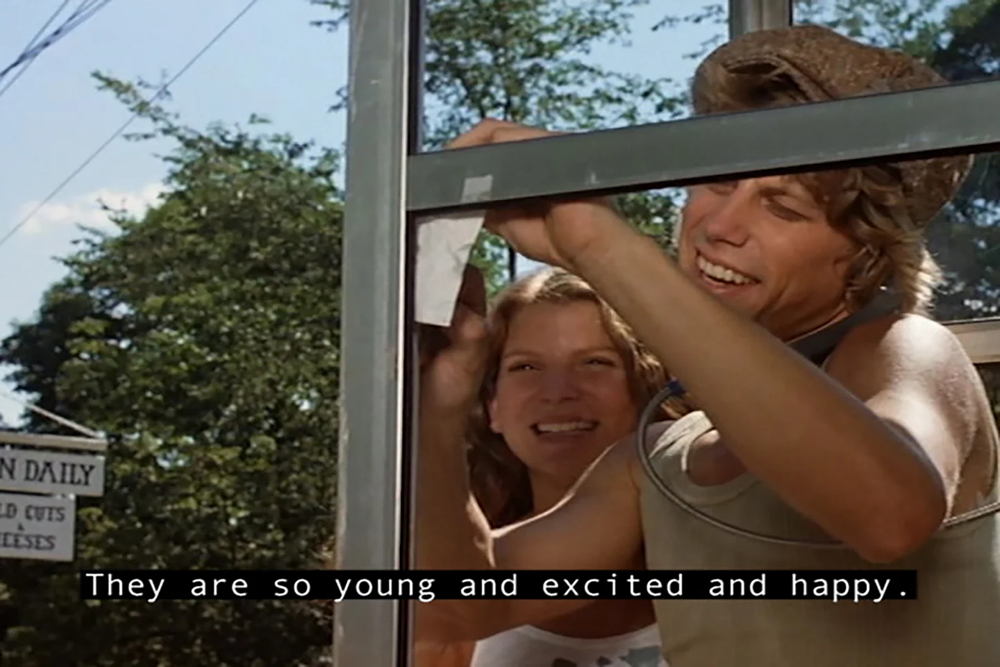 Movie still, close-up of a white man and woman wearing summer clothes in a rural setting looking excited in a phone booth. A caption reads “They are so young and excited and happy.
