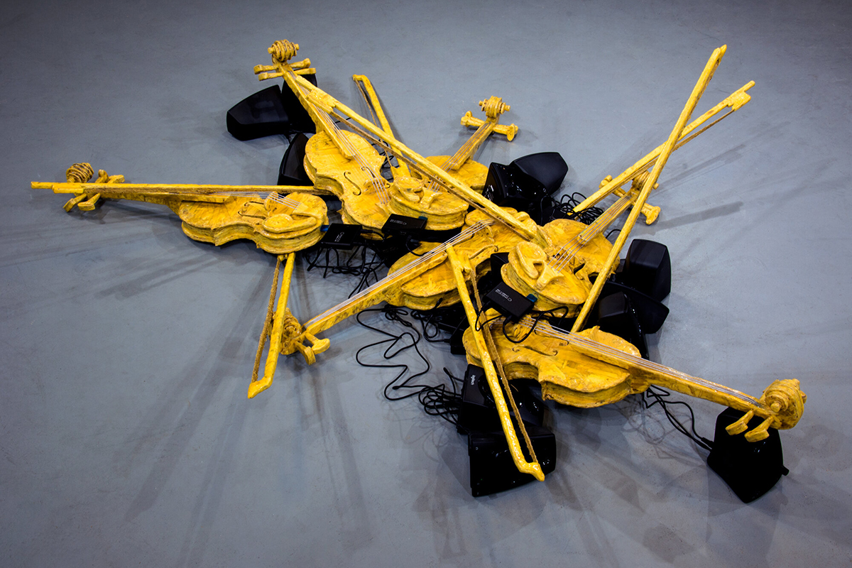  sculpture that looks like a pile of yellow violins and speakers installed on the floor of a gallery